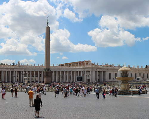 Weitwinkelansicht des Petersplatzes mit Menschen, dem zentralen Obelisken, Brunnen und der Fassade des Petersdoms unter blauem Himmel mit Wolken.