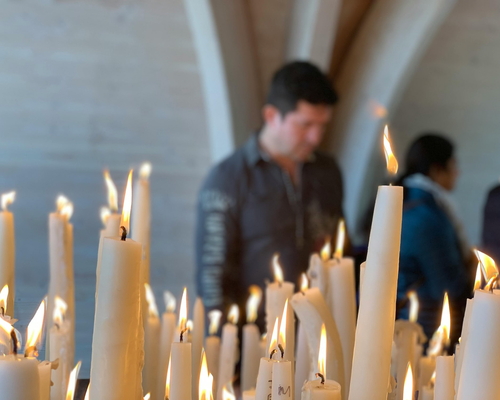 Zahlreiche brennende Kerzen im Vordergrund einer Kapelle in Lourdes, im Hintergrund unscharf zwei betende Personen in stiller Atmosphäre.