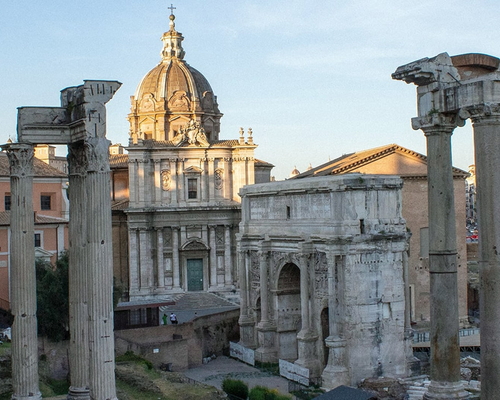 Blick auf die antiken Säulen und Ruinen des Forum Romanum in Rom, mit der barocken Kirche Santi Luca e Martina im Hintergrund bei weichem Abendlicht.